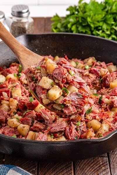 Wooden spoon lifting corned beef hash with potatoes and herbs from a skillet. Salt and pepper shakers, and fresh parsley in the background.