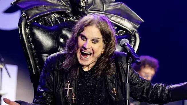 Ozzy Osbourne - a man with long hair and wearing a black shire and black leather coat - sits in a gothic black throne in front of a microphone with his mouth wide open and his arms wide open.