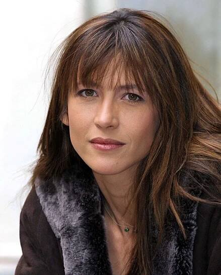 a woman with long brown hair wearing a fur vest and looking at the camera while sitting in front of a window