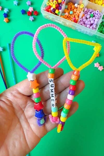 A hand holds three decorated pipe cleaner bubble wands, one pink with letter beads spelling "SUMMER," one yellow with a triangular loop, and one purple with star beads. A bead organizer with pony beads and several colorful pipe cleaners are visible in the background on a green surface.