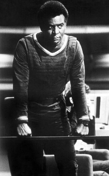 A black and white portrait of Terry Carter in a futuristic military uniform leaning slightly forward with his fists clenched by his side.