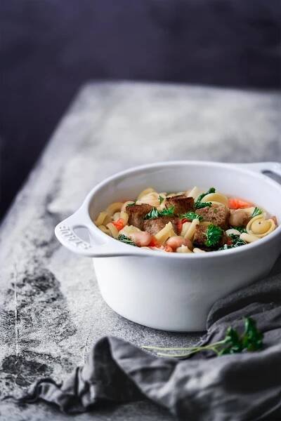 Minestrone aus dem Ofen Oven minestrone Diese Minestrone aus dem Ofen ist die perfekte Schüssel zum Aufwärmen an kalten Herbst-und Wintertagen. Jede Portion wird getoppt mit knusprigen Parmesan-Croutons und frischer Petersilie. Diese Schüssel Minestrone ist eine gemütliche Mahlzeit, bei der man sich einfach wohlfühlen muss. 1,5 Liter Gemüsebrühe 1 Zwiebel, gewürfelt 2 Möhren, gewürfelt ¼ Knollensellerie, gewürfelt 300 g Wirsing, in Streifen geschnitten 1 Lauch, halbiert und in Ringe geschnitten 2 Knoblauchzehen, gehackt 3 Tomaten, gewürfelt 400 g weiße Bohnen, aus der Dose 100 g Pasta ½ Bund Petersilie 200 g Bauernbrot, gewürfelt 25 g Parmesan, gerieben 1 EL Butter Backofen auf 200 Grad Ober-Unterhitze vorheizen. Gemüsebrühe in einen ofenfesten Topf zum Kochen bringen. Zwiebel. Möhren, Sellerie, Wirsing, Lauch, Knoblauch, Tomaten, Bohnen und Pasta in den Topf geben und alles 50 Minuten mit geschlossenem Deckel garen. In der Zwischen die Butter in einer Pfanne erhitzen und die Brotwürfel anbraten, Parmesan darüberstreuen. Suppe mit Croutons und Petersilie servieren.