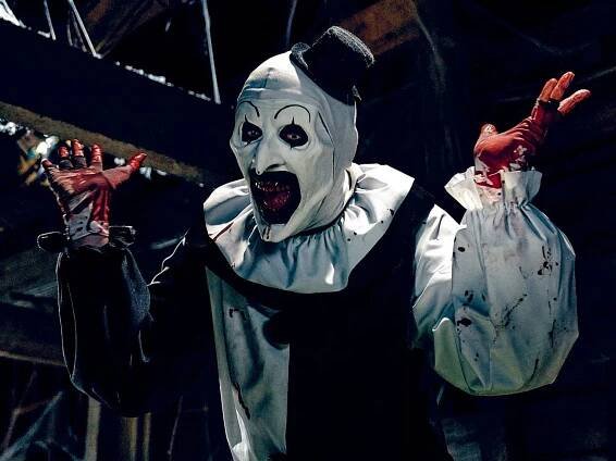 A man in a black and white clown costume with bloody teeth and bloody hands stands in an attic.