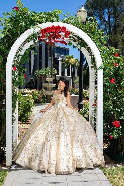 An elegant pose under a white arch decorated with red roses, featuring a sparkly gold ballgown.