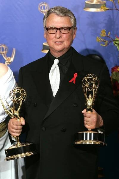 Person in a suit and tie holding two Emmy awards, wearing glasses and a red ribbon on the lapel, in front of a backdrop with Emmy logos
