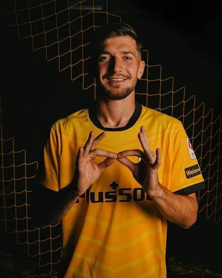 Ein Fussballspieler im gelben Trikot von BSC Young Boys zeigt ein Handzeichen und lächelt in die Kamera, vor einem dunklen Hintergrund mit gelbem Netz.