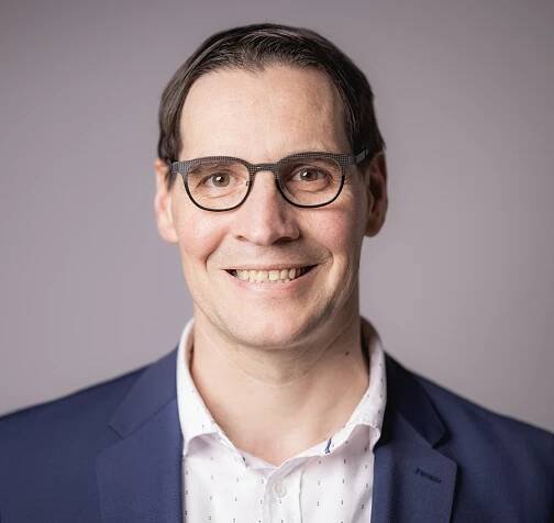 Eine Person mit kurzen Haaren und Brille lächelt, ähnelt Jürgen Müller und trägt ein weißes Hemd und einen dunklen Blazer vor einem schlichten Hintergrund.