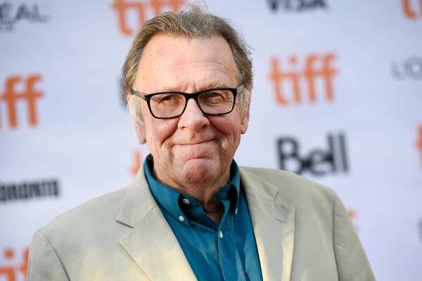 The actor Tom Wilkinson wearing a tan suit jacket, a blue shirt and dark-colored frame glasses. 