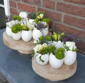 some white eggs with flowers and greenery in them on a blue table next to a brick wall