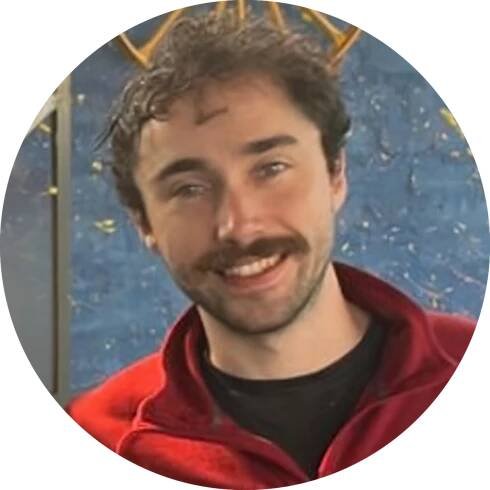 Patrick Ward, smiling with curly hair and a mustache, wearing a red fleece and black shirt, stands in front of a blue artistic background.
