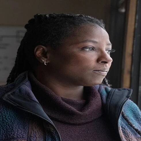 Maria gazing out a window contemplatively, wearing a casual jacket and turtleneck. Background shows a board with faint writing