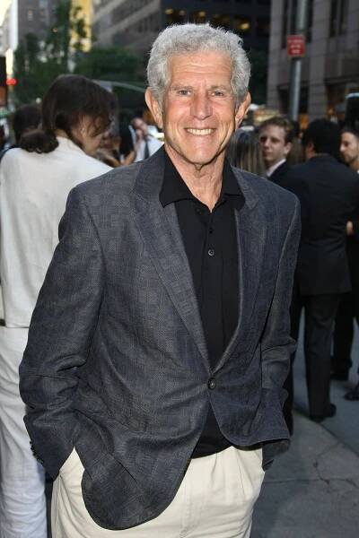 He is standing on a city sidewalk and smiling at the camera. He has short gray hair and wears a charcoal gray sports jacket over a black shirt. 