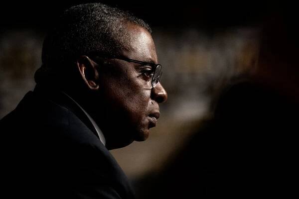 A portrait of Defense Secretary Lloyd J. Austin III. He is visible from the side wearing glasses in low lighting.