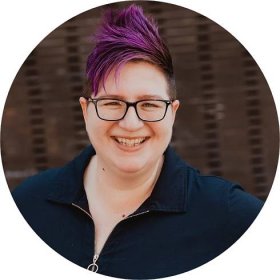 non-binary person with purple hair wearing a navy top with a zip collar and glasses