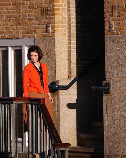 Lea van Acken in der Rolle der Anne Frank aus dem Film Das Tagebuch der Anne Frank. Lea van Ackern, die auf einer Außentreppe vor einem Gebäude steht. Sie trägt eine auffällige, orangefarbene Strickjacke über einem schwarzen, gemusterten Hemd. Dazu kombiniert sie einen braunen Rock. Das Gebäude im Hintergrund hat eine Fassade aus roten Ziegelsteinen. Auf der linken Seite befindet sich die Hausnummer „37“ und auf der rechten Seite die Nummer „39“, was auf ein Wohngebäude hinweist. Die Treppe, auf der sie steht, hat ein Metallgeländer mit Holzverkleidung. Es ist tagsüber, denn die Sonne scheint und wirft deutliche Schatten auf die Treppe und die Wand, wodurch die Szene in warmes Licht getaucht wird.