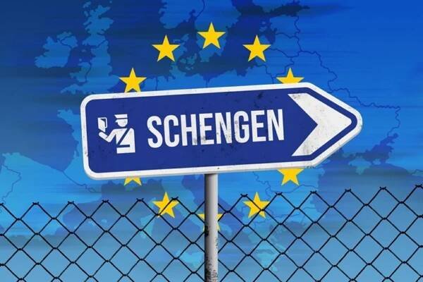 Blue Schengen border sign in front of a map of Europe with yellow EU stars and a wire fence.