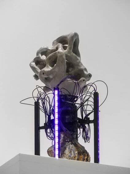 Abstract sculpture composed of a textured gray form atop a rocky base, surrounded by abstract wiring and illuminated by purple lights.