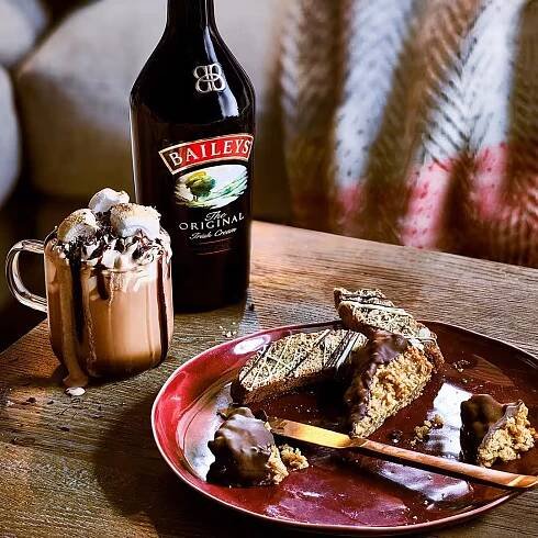 A Baileys hot chocolate sits on a table in a cosy living room. It's topped with marshmallows, cream and chocolate drips.