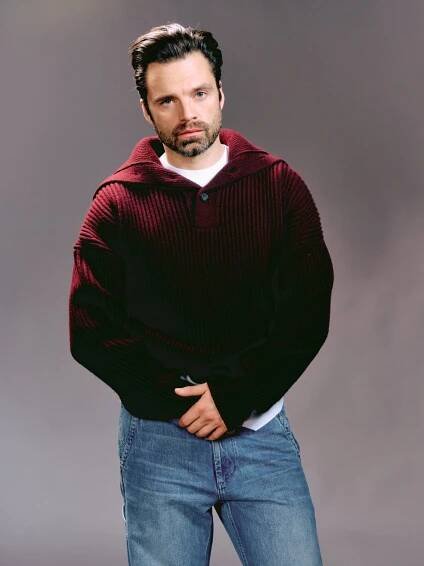 Sebastian Stan is wearing a dark red sweater and bluejeans and posing with his hands crossed in front of him.