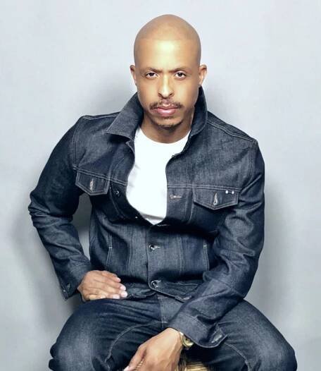 Jamal Smith, in a white t-shirt and denim jacket, gazes at the camera in front of a grey background.