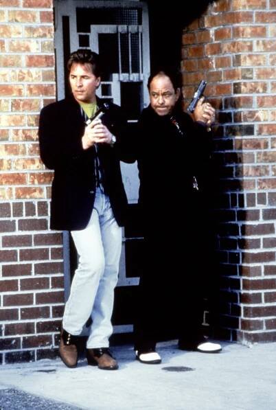 Don Johnson and Cheech Marin in a scene from the original "Nash Bridges" series, holding guns and standing against a brick wall.