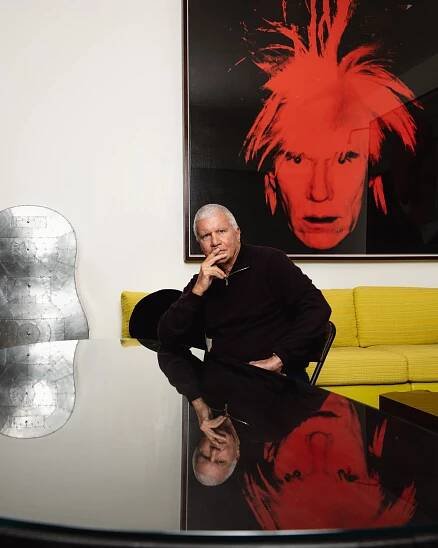 A silver-haired man, Larry Gagosian, sits on a yellow sofa in his home. Above him is a red-on-black silk-screen portrait of Andy Warhol, his hair standing up.