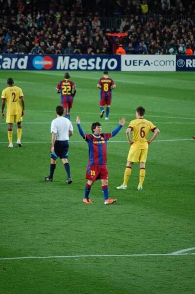 Male football players representing FC Barcelona (red and blue stripes) and Arsenal F.C. (yellow) wearing different colour outfits, while the referee is in a white T-shirt.