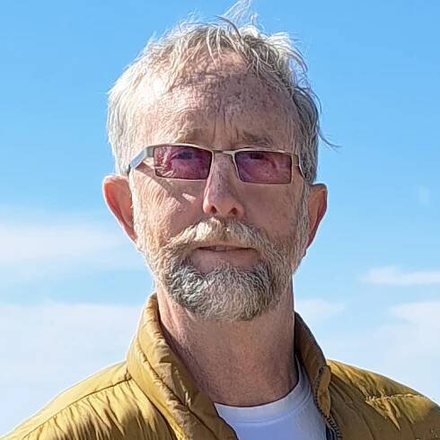 Chris Robinson se tient au bord de l’eau par une journée ensoleillée avec ses lunettes et sa barbe et a l’air très dapper.