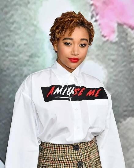 Amandla Stenberg wears a curly pixie cut with a white button-up and plaid skirt