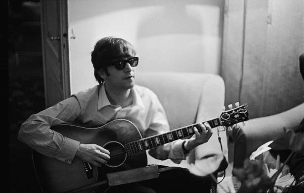 A black and white picture of John Lennon wearing sunglasses and sitting in an arm chair. He plays guitar.