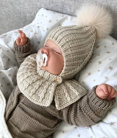 a baby wearing a knitted hat and sweater laying on top of a bed with a pacifier in it's mouth