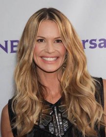 elle macpherson stands in front of an NBCUniversal step and repeat in a black outfit