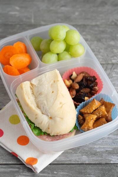 A lunch box with carrots, grapes, a sub, trail mix, and pretzels sitting on a gray table.