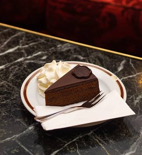 Klassisch angerichtetes Stück der weltberühmten Original Sacher-Torte auf edlem Porzellanteller mit goldener Verzierung. Daneben ein großzügiger Klecks frisch geschlagener Schlagobers, serviert auf einer weißen Serviette mit silberner Gabel.