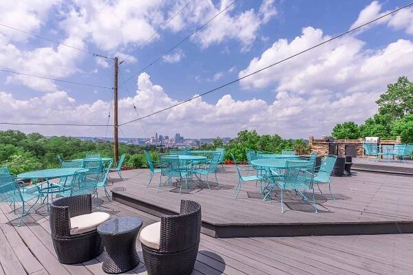 A rooftop patio overlooking a city skyline, featuring turquoise metal tables and chairs arranged on a wooden deck. Lush greenery surrounds the area, and a few black wicker chairs and tables are also present. The sky is bright with fluffy white clouds.
