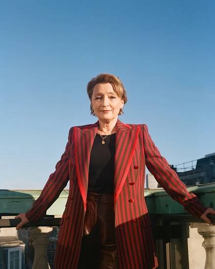 In a portrait, Lesley Manville stands on a balcony, wearing a red striped blazer and against a blue sky.