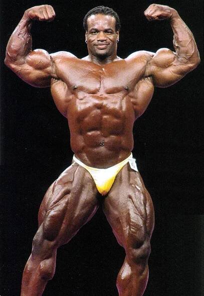 Chris Cormier posing at a competition, showing his large quads, ripped abs and large bulging biceps
