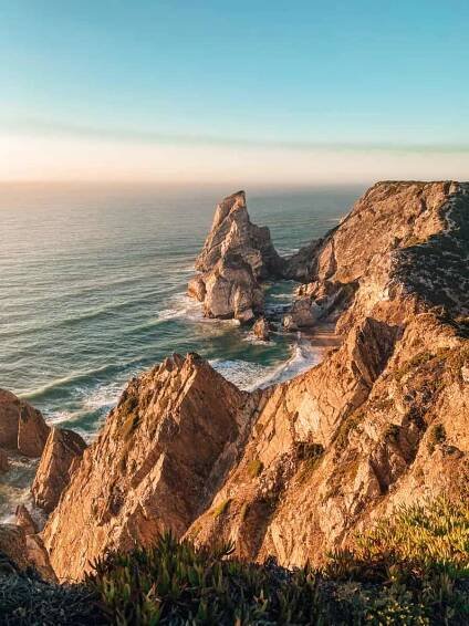 A breathtaking sunset view over Ursa Beach, Portugal, with golden cliffs illuminated by the warm glow of the fading sunlight. The towering limestone sea stacks rise dramatically from the Atlantic Ocean, as waves gently lap against the secluded sandy shore. One of Portugal’s most stunning hidden beaches near Cabo da Roca, known for its rugged beauty and remote charm.
