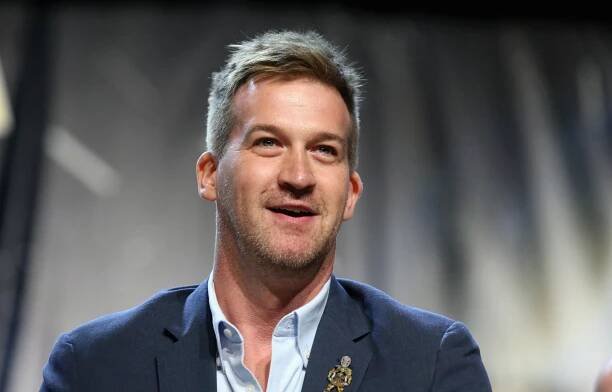 A man with blond hair and stubble, wearing a blue suit with no tie and a Star Trek pendant.