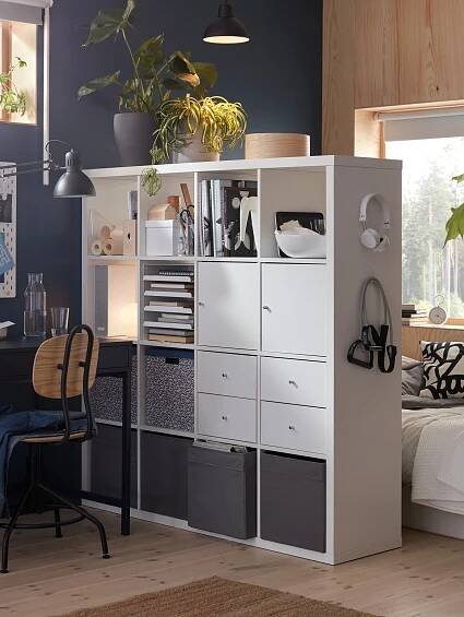Ein weißes KALLAX Regal in 4x4 steht als Raumteiler zwischen einem Schlafzimmer und Arbeitszimmerbereich. In dem Regal sind 2 weiße KALLAX Türen und 4 weiße KALLAX Schubladen sowie diverse Boxen, Fächer sowie Bücher untergebracht.