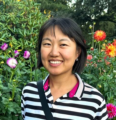 portrait photo of a smiling east asian woman with chin-length black hair, wearing a black and white striped shirt, in front of a background of trees and dahlias