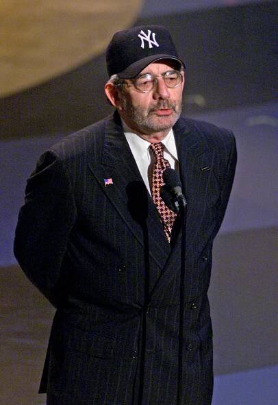A bearded, middle-aged Gary Smith, wearing glasses, a baseball cap, a suit and a tie, stands behind a microphone, his hands behind his back.