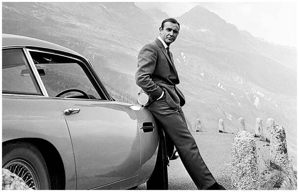 Černobílý plakát s Seanem Connerym jako Jamesem Bondem opřeným o auto Aston Martin na pozadí hor.