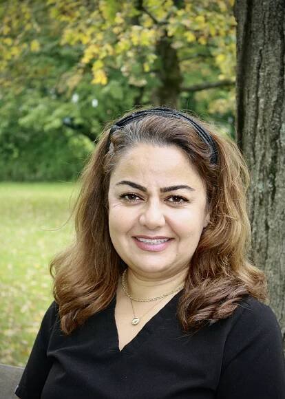 A smiling woman with dark hair wearing a black top and a headband, posing for a portrait against an autumnal backdrop.