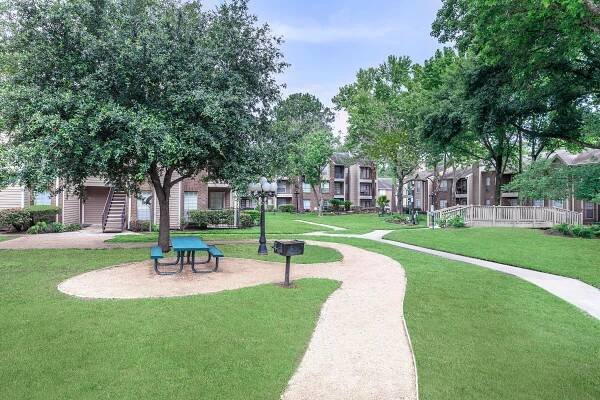 A landscaped courtyard featuring green grass, trees, and walking paths. There are picnic tables and lamp posts, with residential buildings visible in the background. The scene is bright and inviting, ideal for outdoor gatherings or leisurely strolls.