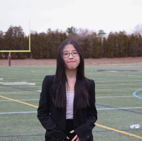 Jennifer with long dark hair is smiling at the camera while wearing glasses on a sports field.