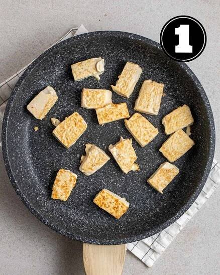 Pan-frying silken tofu cubes in a non-stick pan on a white folded checkered teacloth.