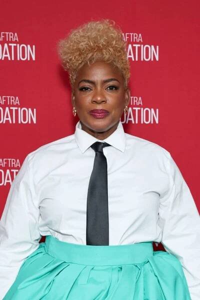 Person in a crisp white shirt, black tie, and teal skirt at a red carpet event, standing in front of a screen with "SAG-AFTRA Foundation" text