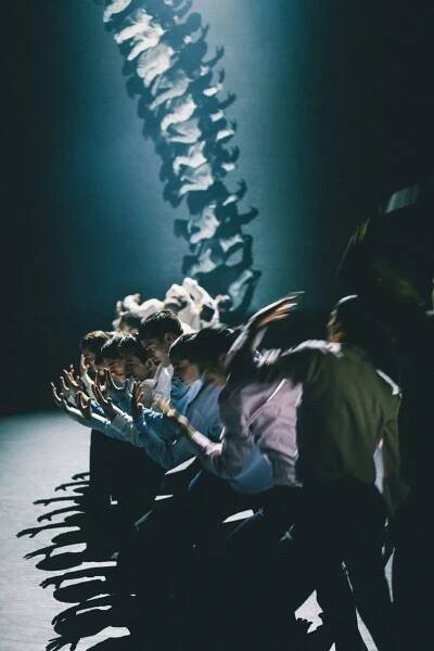 In front of a projection of a spinal cord, a curved line of dancers use their bodies to create shadows on the floor that echo the structure of a spine.