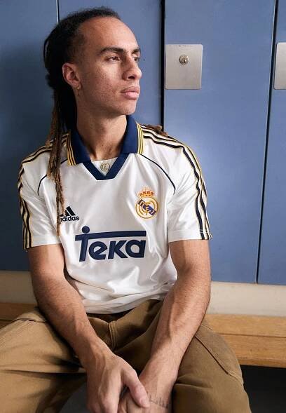 Bílý dres adidas s modrým límečkem a žlutými akcenty, obsahuje logo 'Teka' a znak Real Madrid. Nosený s béžovými kalhotami, sedící proti skříňkám.